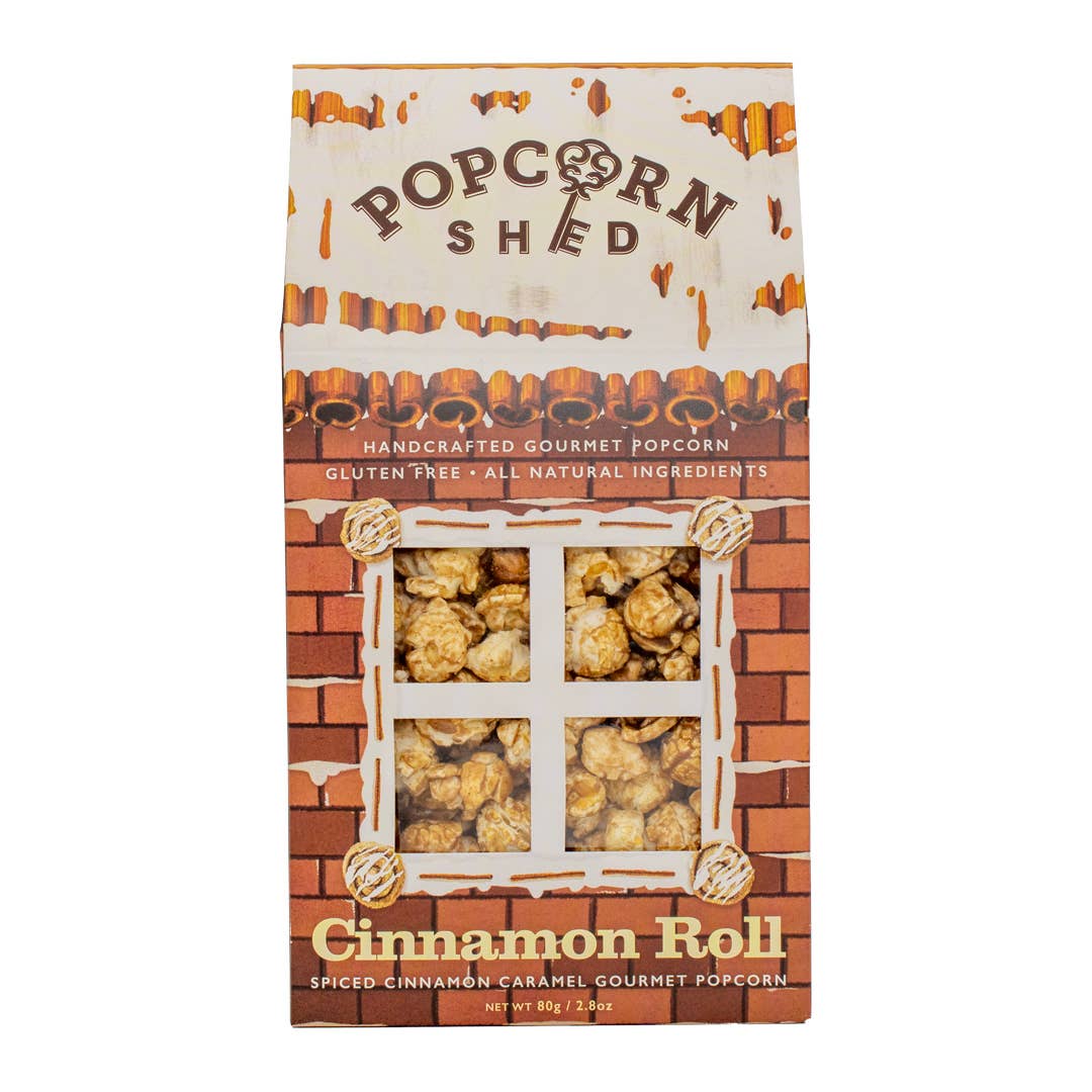 A box of Cinnamon Roll Popcorn Shed, handcrafted in the UK and featuring a charming brick house design with a window displaying delicious caramel popcorn, highlights that it is gluten free, all natural, and boasts a spiced cinnamon caramel flavor.