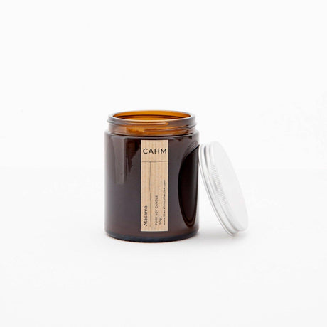 A minimalist brown glass jar labeled "CAHM," crafted from eco-friendly materials, contains an Atacama Amber Jar Candle. Its lid sits beside it, and a beige label highlights the natural wax inside against a plain background.