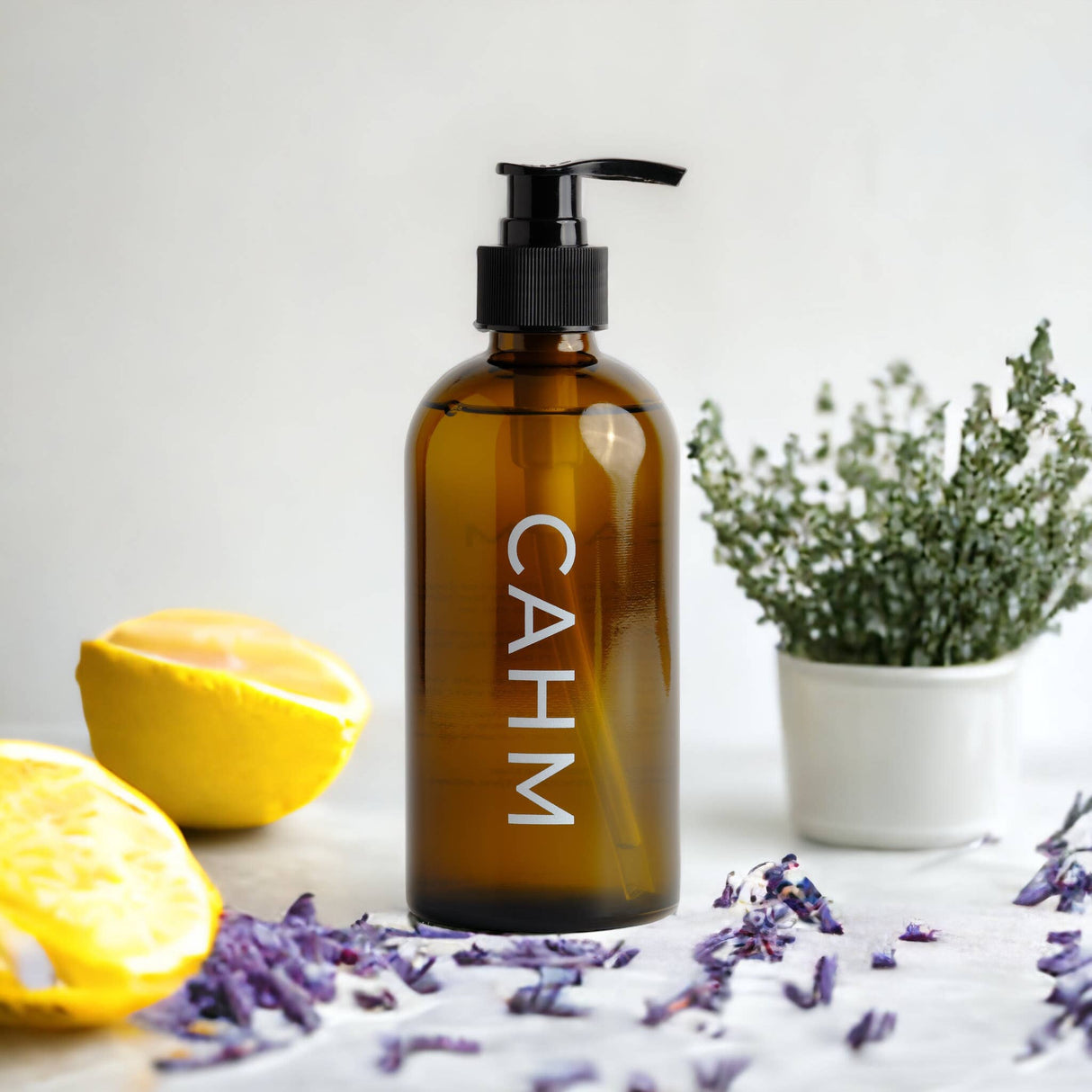 A brown bottle with a black pump labeled "CAHM" features the zesty scent of Amalfi Coast Hand Wash. It's set on a white surface with purple dried flowers, half a lemon, and a small potted plant against a softly blurred background.
