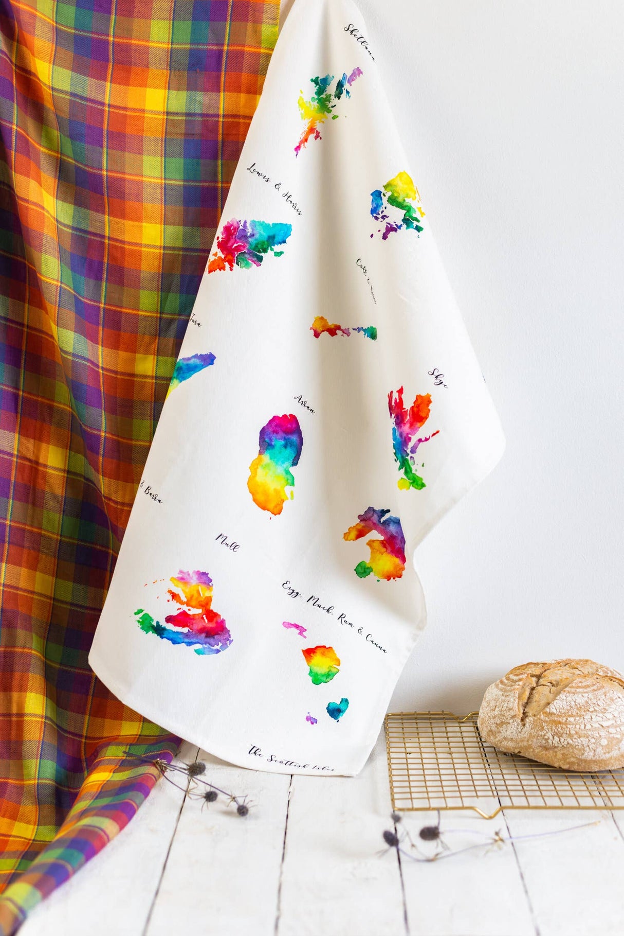 A colorful tartan fabric hangs beside a Scottish Islands Ceramic Jar from Sarah Leask Studio, decorated with vibrant, watercolor-style island shapes. A loaf of bread and a wire cooling rack rest on a rustic white surface.