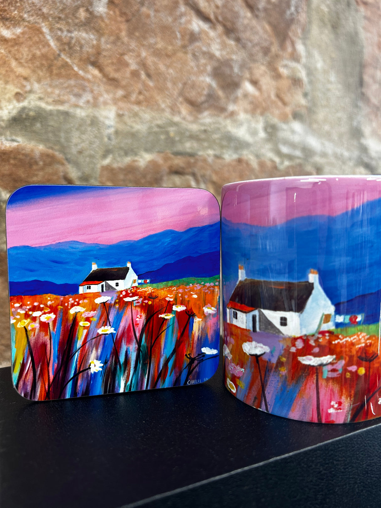 The Pink Sky Mug showcases white cottages, colorful wildflowers, and mountains under a vivid pink and blue sky, capturing the charm of a Scottish landscape with a stone wall backdrop.