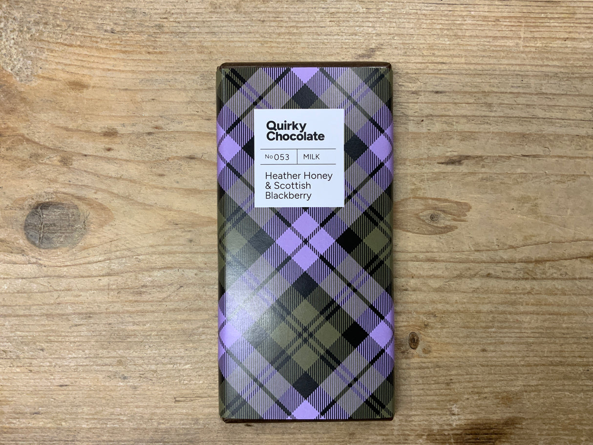 The Heather Honey & Scottish BlackBerry Milk Chocolate bar comes in tartan-patterned packaging of purple, black, and olive green stripes. Made with heather honey and Scottish blackberries, it is shown on a wooden surface.