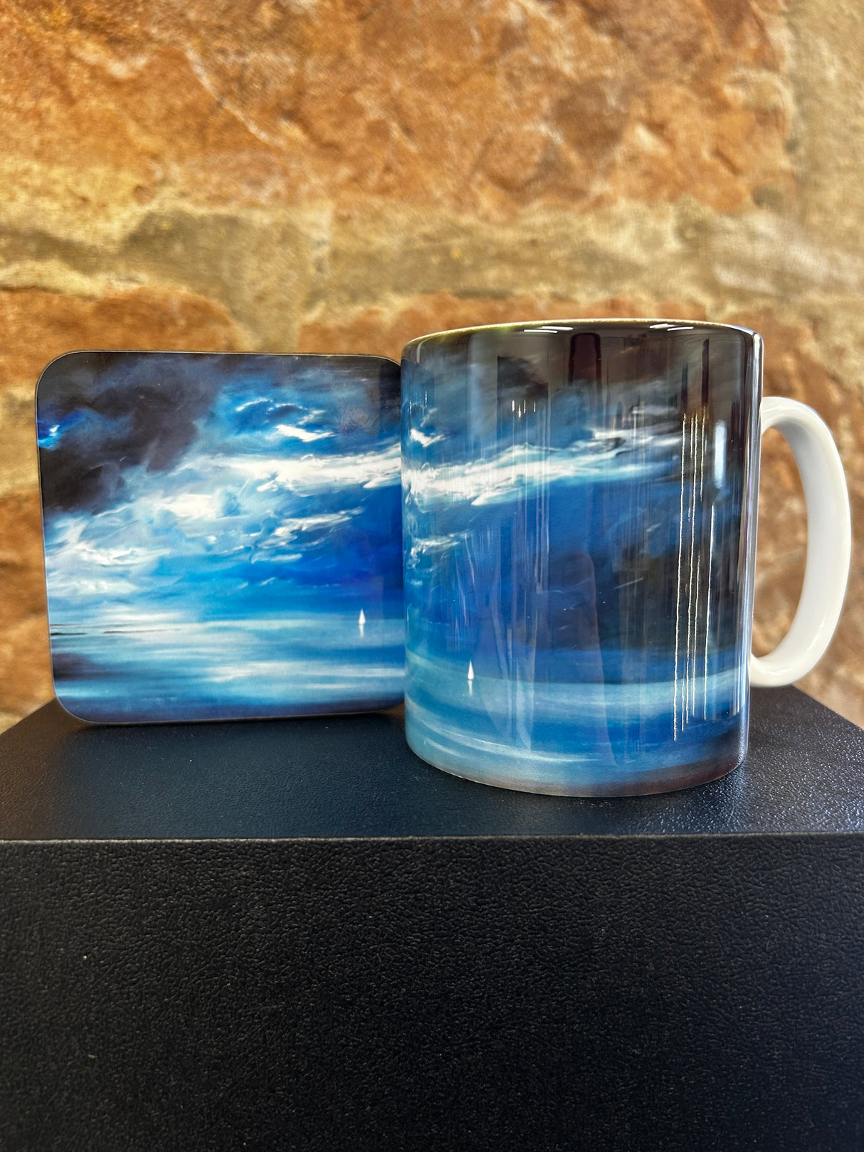 The Blue Yonder Mug and its matching coaster, both with a vibrant blue and white abstract cloud design, are displayed against a textured brown stone background.
