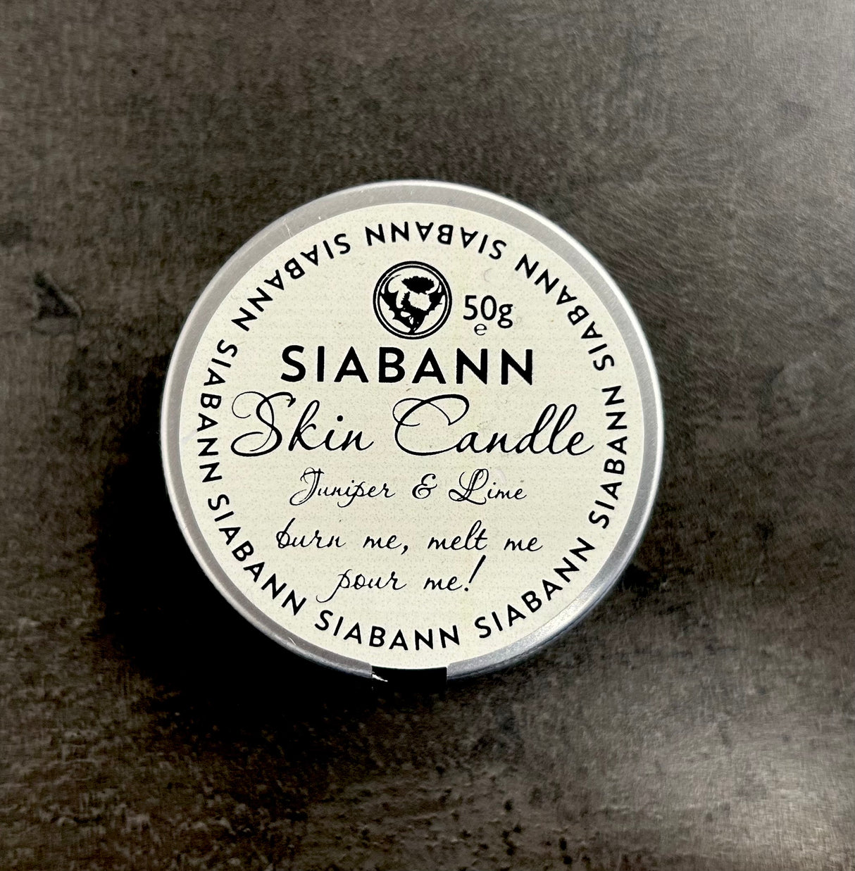 A 50g Siabann Juniper & Lime Skin Candle rests on a dark surface. Its label reads, "burn me, melt me, pour me!"—showing it doubles as a nourishing massage oil for your skin.