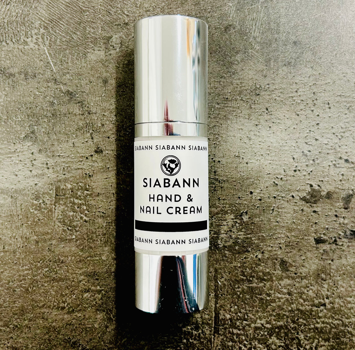 A silver cylindrical bottle of Siabann Hand & Nail Cream 30ml stands upright on a textured gray surface. Its white label with black text features Sweet Almond oil and Juniper & Lime, along with a small logo above the brand name.