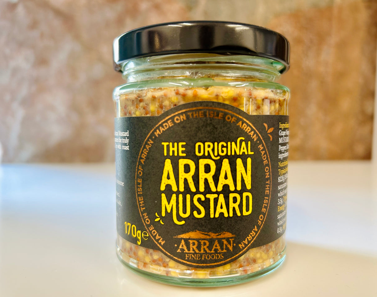 A 170g glass jar of "Original Arran Mustard" by Arran Fine Foods, showcasing its Isle of Arran origin on the label, is situated on a light surface with a softly blurred backdrop.