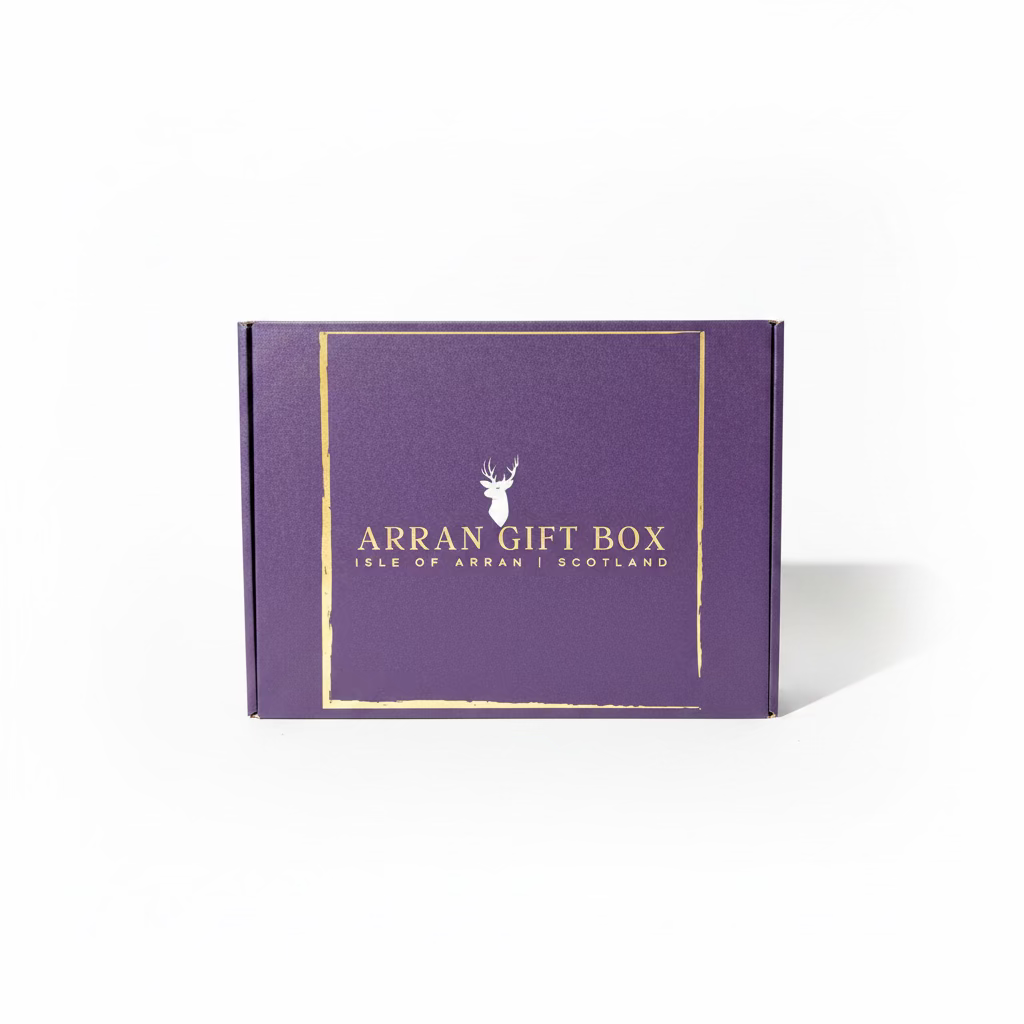 A box with a purple shade and golden text reading "Arran Gift Box - Medium Size Gift Box, Isle of Arran, Scotland" rests on rocky terrain near the water's edge, promising a delightful experience of Scottish delicacies. The backdrop features a serene lake, distant mountains, and a bright blue sky.