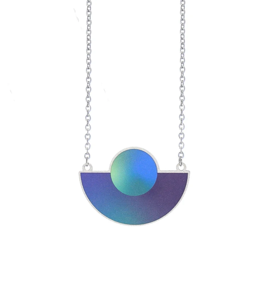 The Cosmic Moon Necklace Space Blue boasts a modern, abstract design with a silver chain and a geometric pendant that features a semi-circle in purple and green hues and an overlapping circle on top.