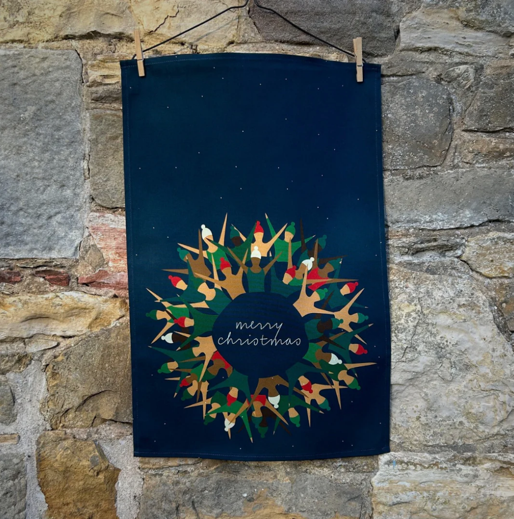 The Festive Swim Tea Towel, crafted from premium cotton, showcases a stylized wreath design in green and red with the wording "merry christmas." It hangs against a rustic stone wall, and its dark blue background enriches the festive theme.