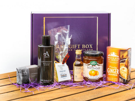 The Original Lochranza (10 Year Old Malt) Arran Gift Box presents a charming selection: Lochranza bath and shower gel, Arran whisky, marmalade, ginger rounds, and chocolate. Set against a wooden backdrop with purple shredded paper, this gift box perfectly embodies the luxurious essence of Arran indulgence and skincare elegance.