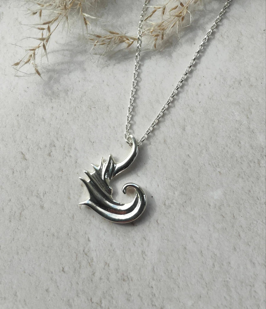 The "Tide Keeper" Necklace is a sterling silver pendant featuring a stylized dragon or wave, shown on a light textured surface with dried grasses in the background.