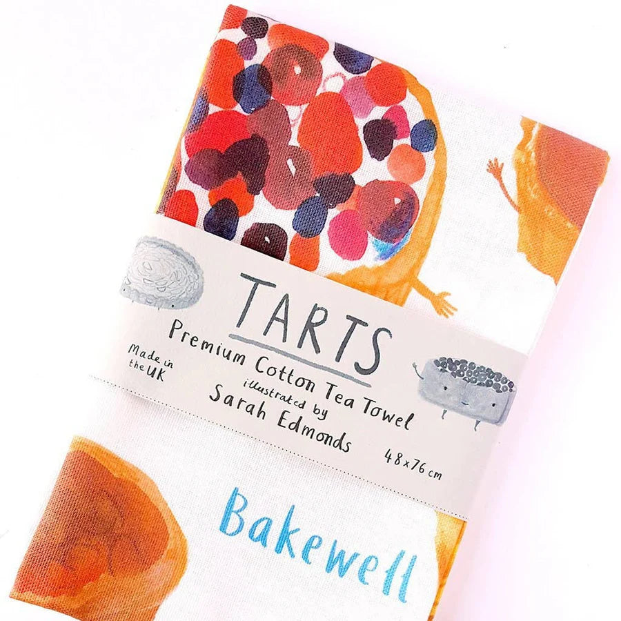 A folded tea towel featuring colorful tart illustrations, labeled "TARTS Premium Cotton Tea Towel, illustrated by Sarah Edmonds," with "Bakewell" written on the visible part of the fabric.