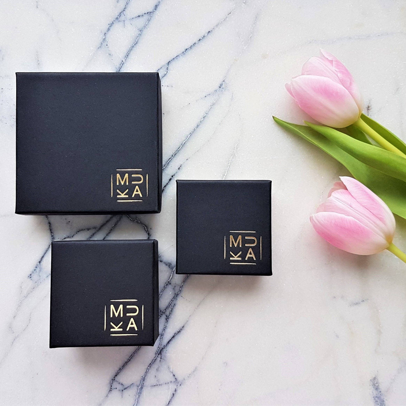 Three black square gift boxes with gold logos are arranged on a white marble surface next to two pink tulips, creating a minimalist and elegant composition.