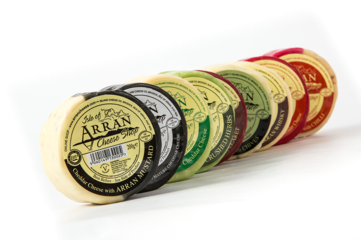A row of colorful cheese rounds from the Isle of Arran Cheese Shop. The flavors include Arran Mustard, Oak Smoked, Chive, Crushed Herbs, and Whisky. Each is wrapped in different colored packaging.