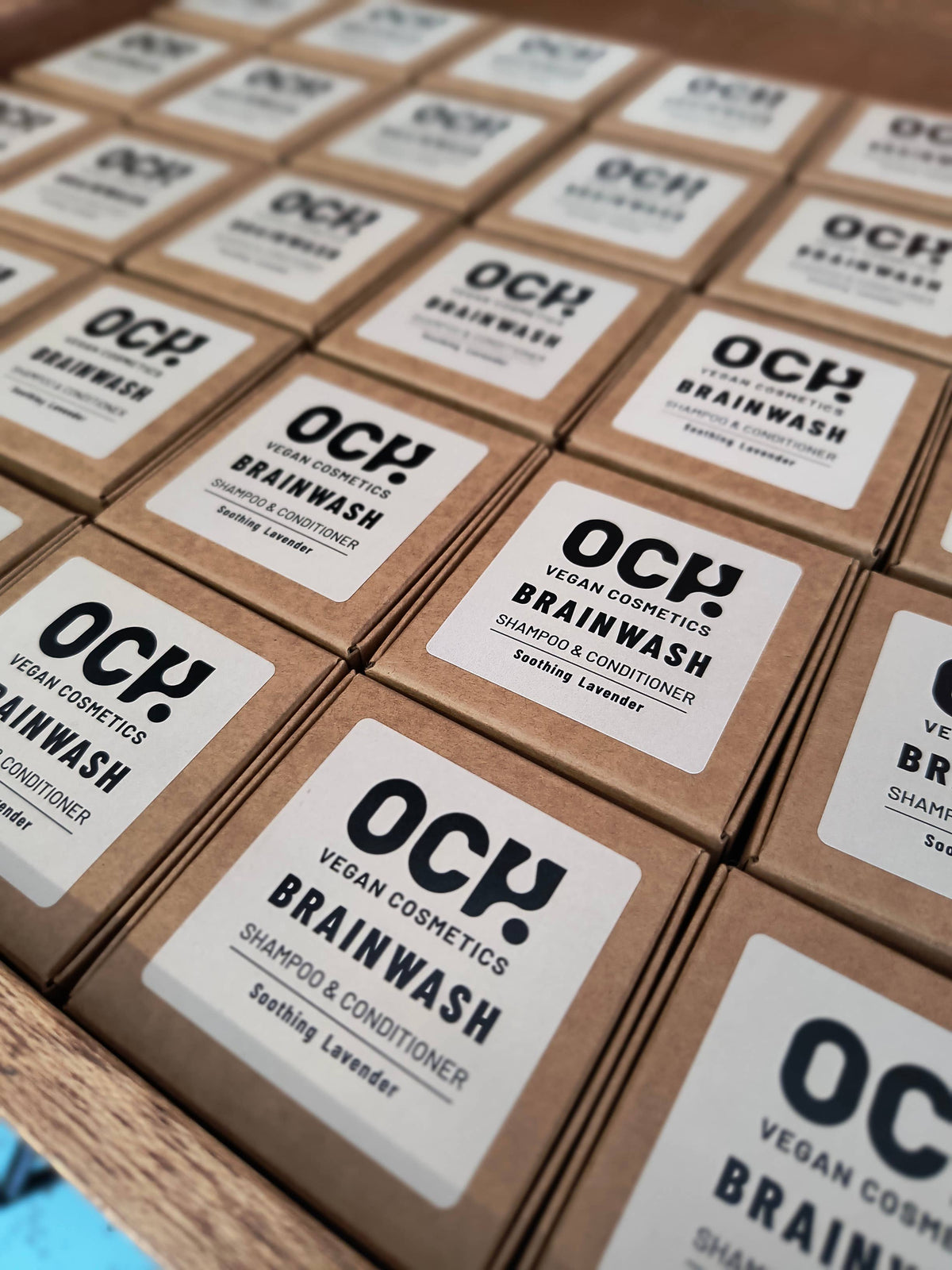 Rows of brown cardboard boxes labeled “OCY! Vegan Cosmetics Brainwash Shampoo & Conditioner Soothing Lavender” are neatly arranged on a wooden shelf.