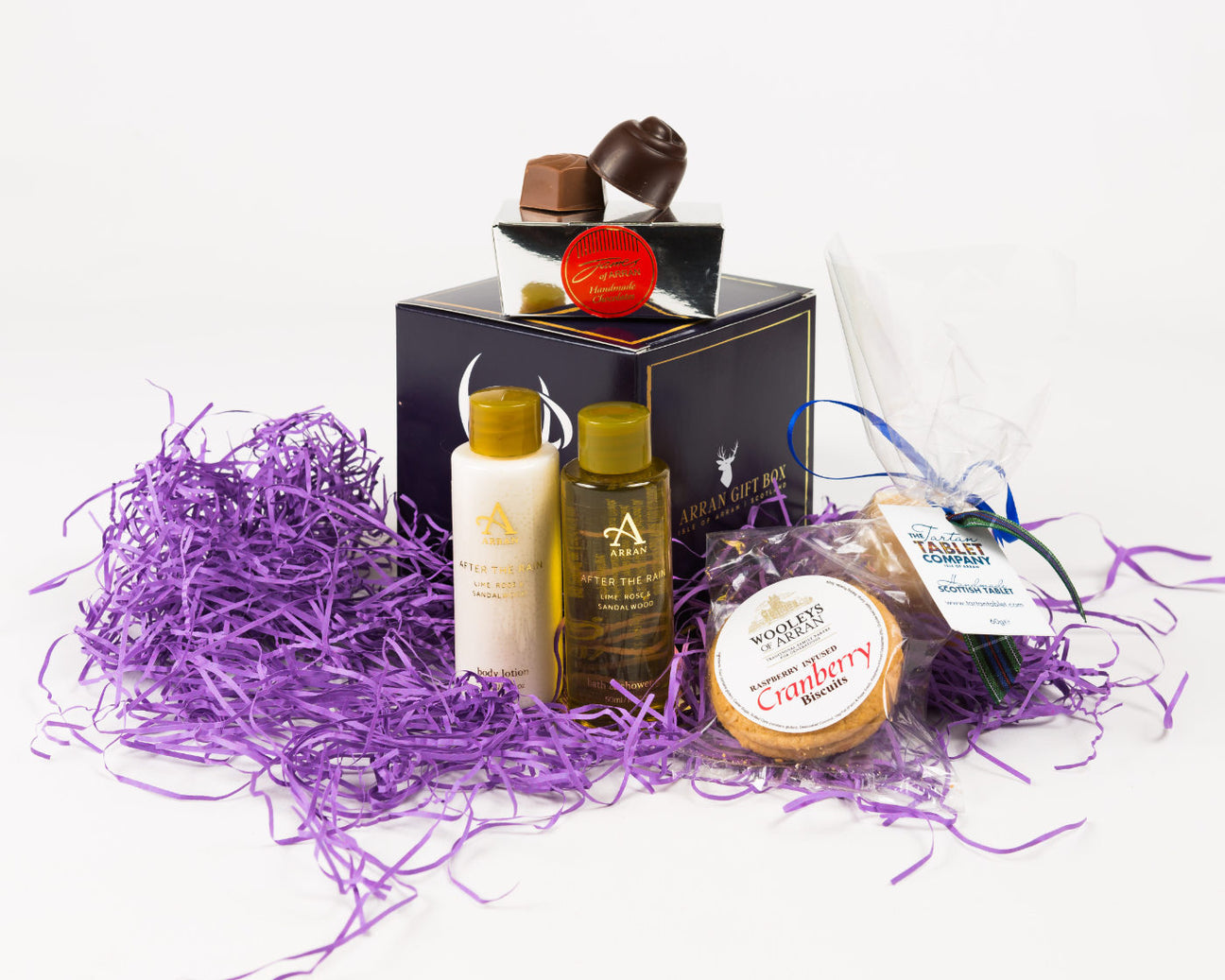 Gift box with chocolates, two bottles of toiletries, shortbread, and a food item wrapped in clear packaging on purple shredded paper.
