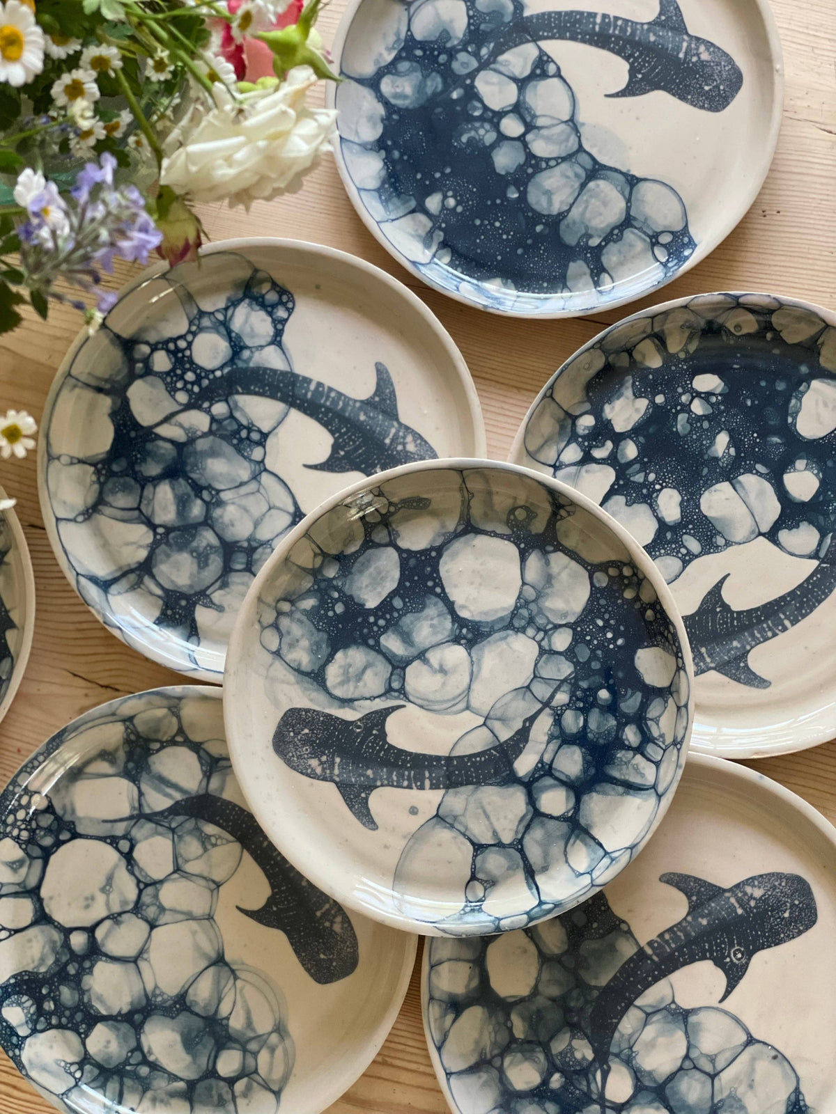 Ceramic plates with blue whale designs rest on a wooden surface. The intricate patterns resemble ocean waves. A small floral arrangement with daisies and greenery is visible in the corner.