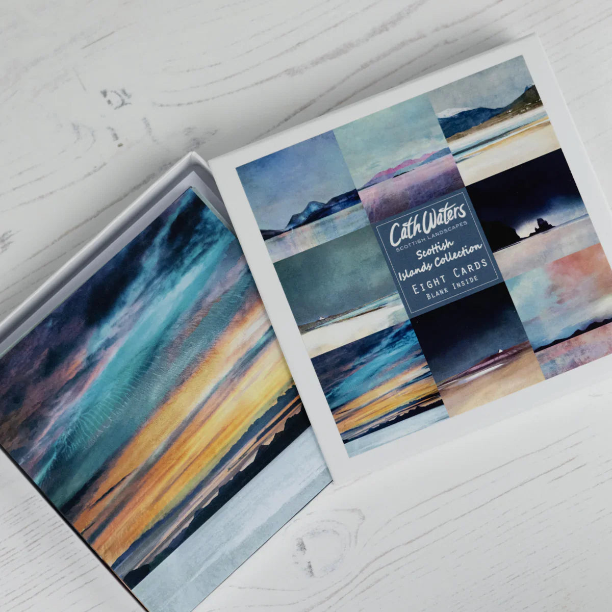A box set titled "Cath Waters Scottish Islands Collection" with eight art cards, each featuring colorful landscape paintings of skies, sea, and islands, displayed on a white wooden surface.