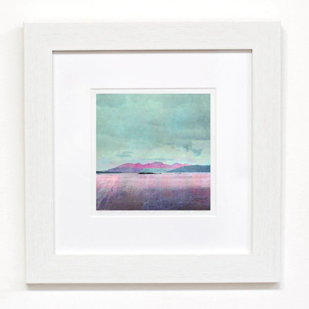 Framed artwork featuring a pastel-colored seascape with pink and purple mountains under a cloudy sky, surrounded by a white mat and white frame on a white wall.