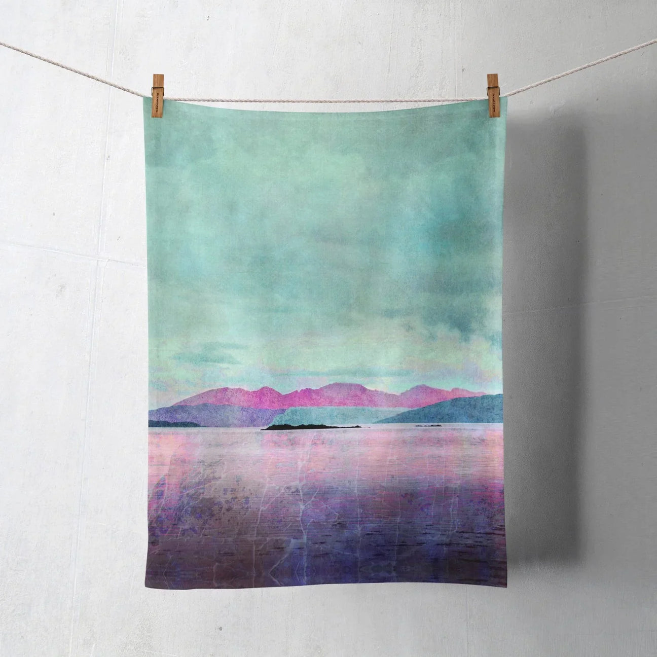 A pastel-colored tea towel with a landscape design featuring a calm lake, distant mountains in pink and blue tones, and a cloudy sky, hanging on a string with clothespins.