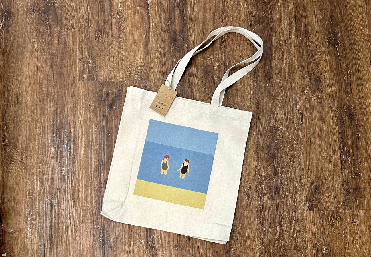 A beige tote bag with two long handles lies on a wooden floor. The bag features a minimalist illustration of two people standing on a yellow and blue background, resembling a beach scene.
