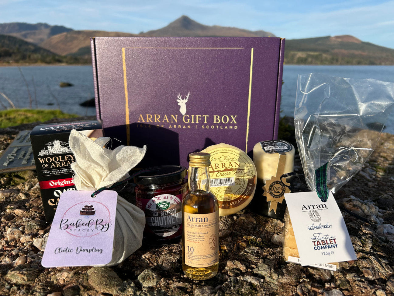 A gift box labeled "Arran Gift Box" is surrounded by various local Scottish products, including cheese, chutney, whisky, and oatcakes, set on a rocky surface with mountains and a lake in the background.