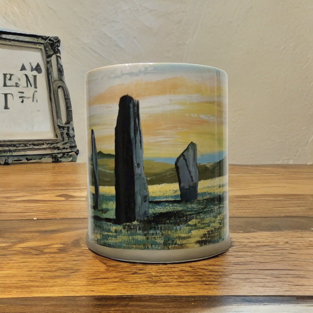 A ceramic mug with an illustration of standing stones in a grassy landscape, set against a yellow and gray sky; a framed picture sits in the background on a wooden surface.