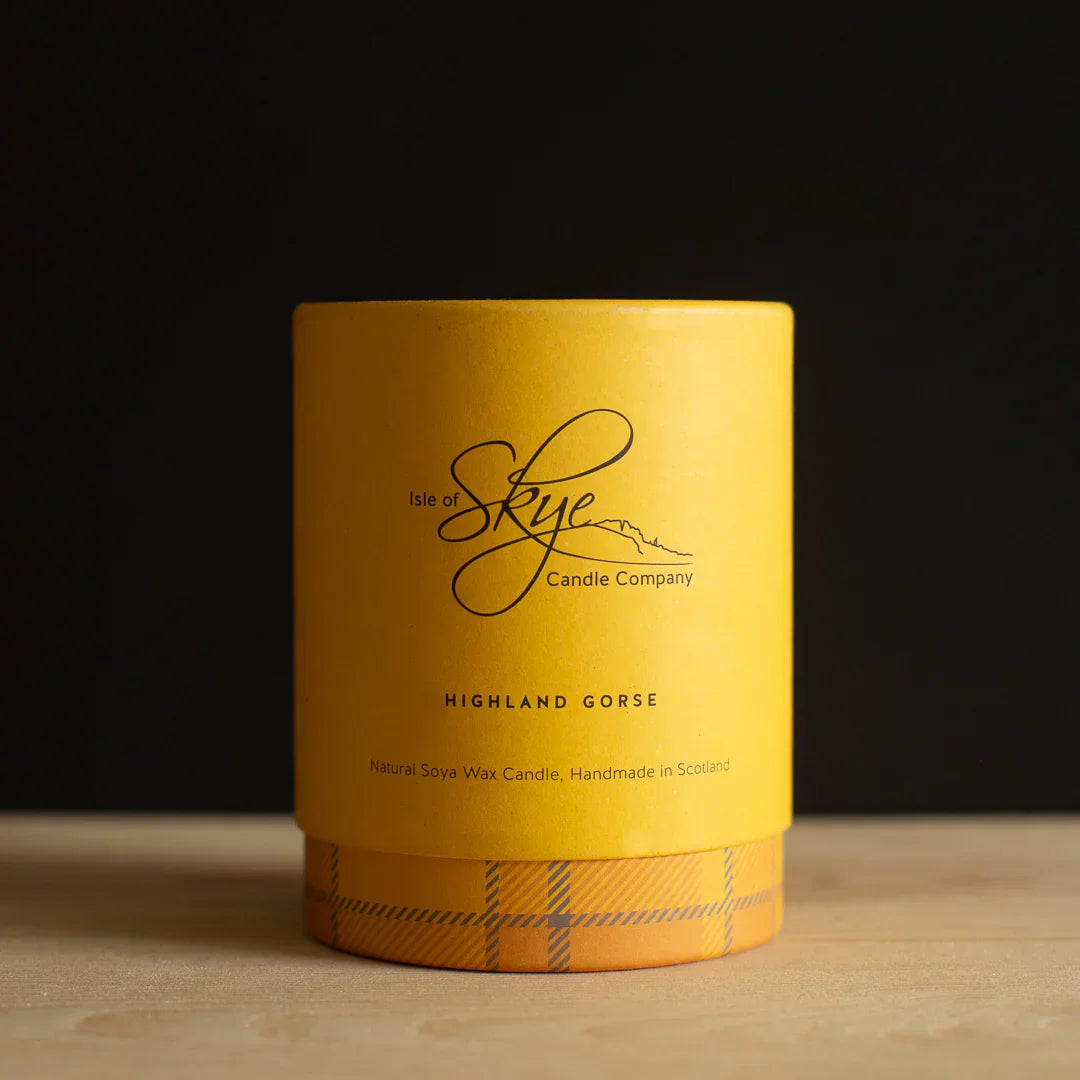 A yellow candle in a cylindrical container labeled "Isle of Skye Candle Company, Highland Gorse" sits on a wooden surface against a black background.