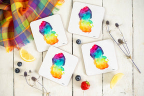 Four square Arran Watercolour Map Coasters with colorful abstract art are arranged on a white wooden table, alongside blueberries, a strawberry, lemon wedges, blue thistles, and a plaid cloth for Scottish home decor charm.