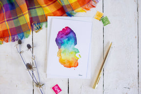 The Arran Map Greetings Card, featuring a colorful watercolor map of the Isle of Arran, is shown on a white wooden surface beside a gold pen, dried flowers, stamps, and a vibrant plaid cloth.