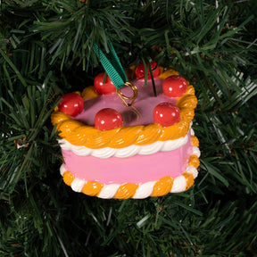 A Heart, Heart Cake ornament with pink frosting, white trim, and red cherries hanging on a Christmas tree.