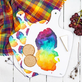 The Arran Chopping Board Small, featuring vibrant watercolor art inspired by Scottish homeware, sits on a white surface with a plaid cloth, two round crackers, cheese, and grapes.