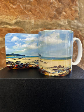 The Whiting Bay to Holy Isle Mug, featuring a painted beach scene with sand, rocks, blue sky, and clouds, is displayed against a textured brown stone background.
