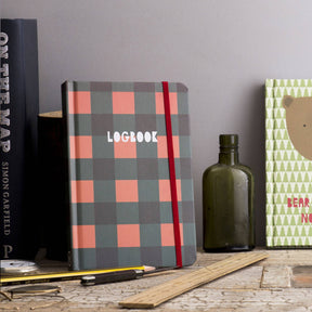 A Lumberjack Notebook featuring a plaid A6 cover with a red elastic band stands upright on a desk, surrounded by a ruler, pen, pencil, green glass bottle, and books—including one with a bear illustration.