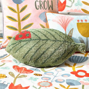 The Little Nature Leaf Knitted Kids Cushion Green, with a red ladybird design, sits on a floral bedspread among colorful pillows decorated with flower prints and "GROW," creating charming nature-themed kids’ décor.