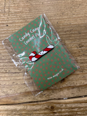 A small Candy Cane Enamel Pin featuring a smiling candy cane is attached to a green card with red stars, labeled "Candy Cane Enamel Pin," wrapped in clear plastic, and placed on a wooden surface.