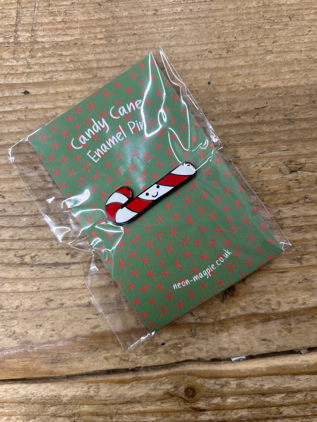 A small Candy Cane Enamel Pin featuring a smiling candy cane is attached to a green card with red stars, labeled "Candy Cane Enamel Pin," wrapped in clear plastic, and placed on a wooden surface.