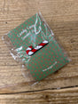 A small Candy Cane Enamel Pin featuring a smiling candy cane is attached to a green card with red stars, labeled "Candy Cane Enamel Pin," wrapped in clear plastic, and placed on a wooden surface.
