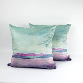 Two Arran Velvet Cushions with a soft watercolor-style landscape in pastel green, purple, and pink, featuring mountains and water, showcase contemporary Scottish design against a plain white background.