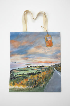 The Road to Kildonan Tote Bag features cream handles and a scenic landscape painting of a coastal road, fields, and a vibrant sunset sky, with a brown tag on one handle. The bag is shown on a white background.