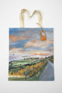 The Road to Kildonan Tote Bag features cream handles and a scenic landscape painting of a coastal road, fields, and a vibrant sunset sky, with a brown tag on one handle. The bag is shown on a white background.