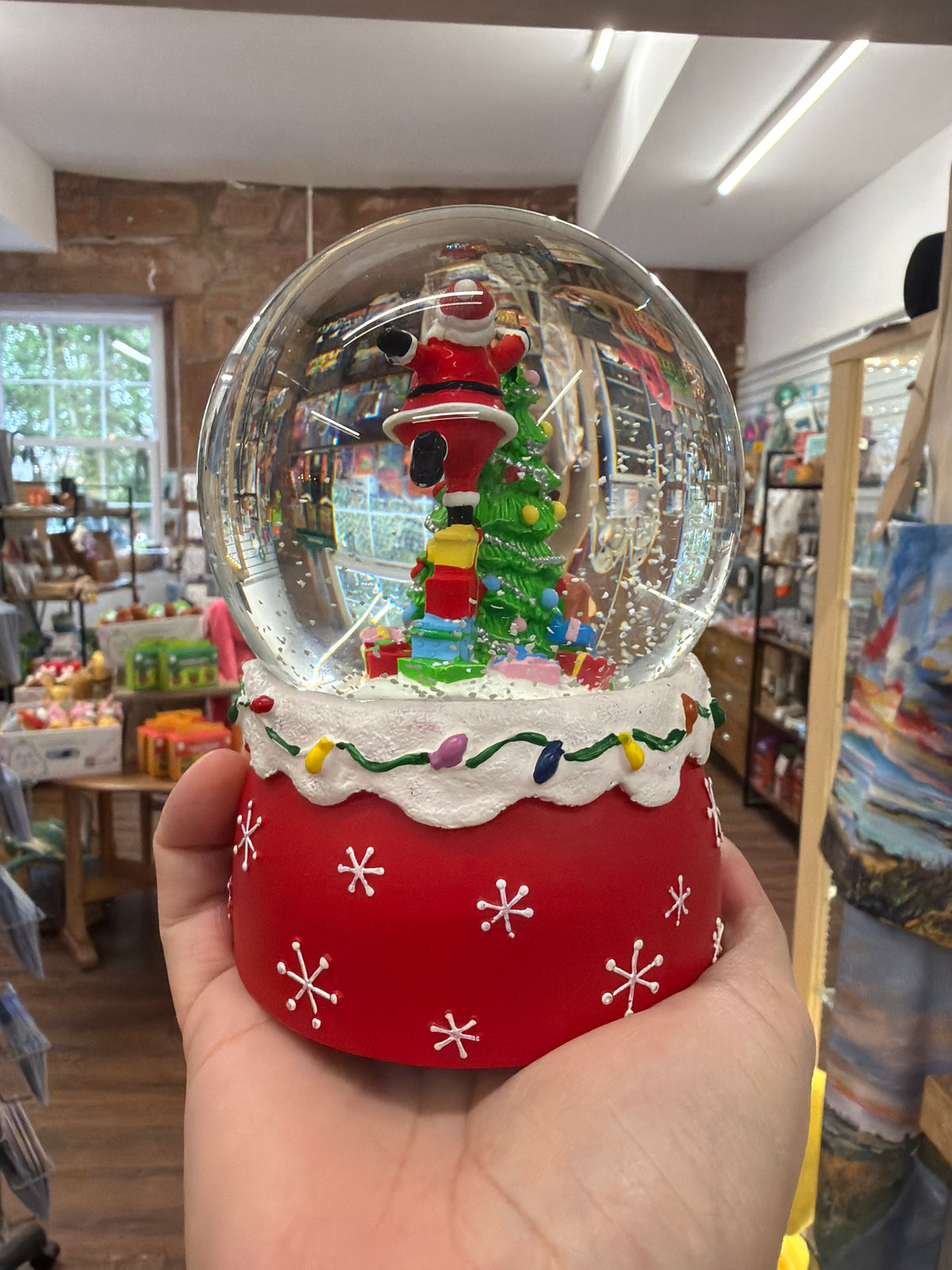 A person holds the Musical Snow Globe, 15cm - Santa & Tree/Red, which features a decorated Christmas tree, presents, and Santa Claus inside. In the background, store shelves display colorful items.