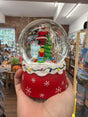 A person holds the Musical Snow Globe, 15cm - Santa & Tree/Red, which features a decorated Christmas tree, presents, and Santa Claus inside. In the background, store shelves display colorful items.