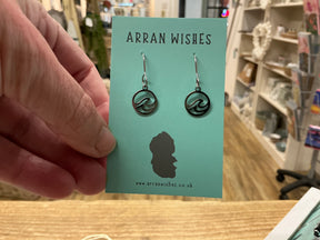 A hand displays an "Arran Wishes Earrings - Hypoallergenic" turquoise card featuring cute wave design earrings. Behind, a cozy shop with wooden floors and shelves showcases various crafts.