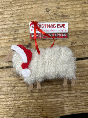 The Christmas Ewe Hanging Decoration, a woolly sheep ornament with a red Santa hat and "Handmade Christmas Hanging" tag, rests on wood—a perfect Scottish gift, handmade in Scotland.