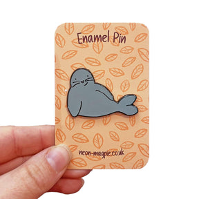 A hand holds a card displaying the Seal Enamel Pin, featuring a smiling gray seal. The card shows a leaf pattern, "Enamel Pin" at the top, and "neon-magpie.co.uk" at the bottom—the perfect cute accessory for any pin collection.