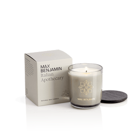A Max Benjamin – Italian Apothecary Natural Candle, hand-poured in Ireland with natural wax, sits in a glass holder with a black lid beside its cream-colored box on a white background.