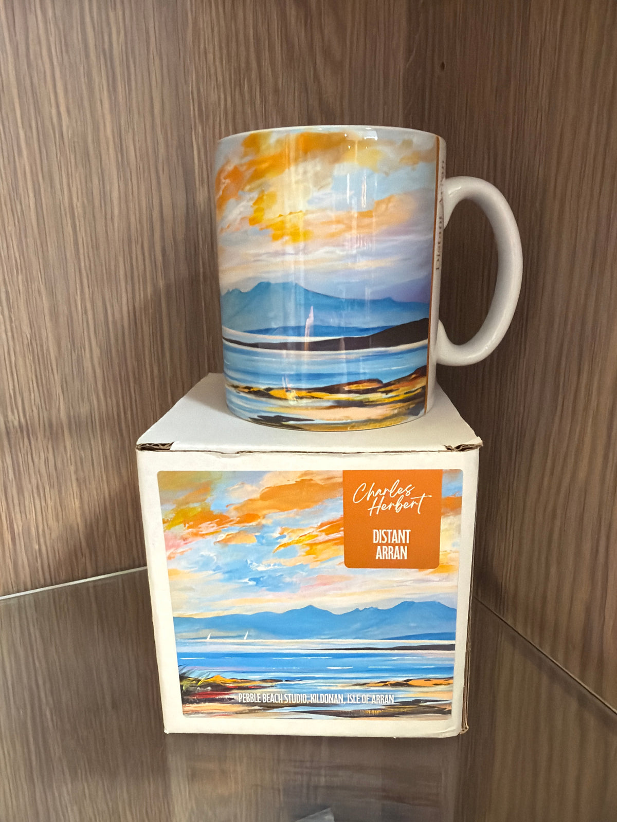 The Distant Arran Mug, adorned with Charles Herbert’s vibrant landscape of mountains and water, is perched on its matching box labeled “Distant Arran” and beautifully showcased against a wooden shelf.