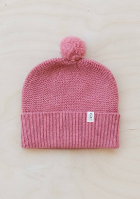 The Merino Wool Baby Beanie in Rose has a pom-pom, folded brim, and soft Merino wool. It features a small white front tag and is shown on light wood.