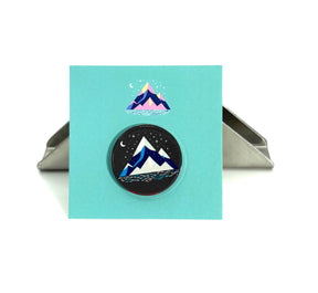 The Arran Wishes Grey Mountain Enamel Pin, featuring a snow-capped mountain scene with a crescent moon and stars, is displayed on a turquoise card with a matching skyline background and held upright by a metal stand.
